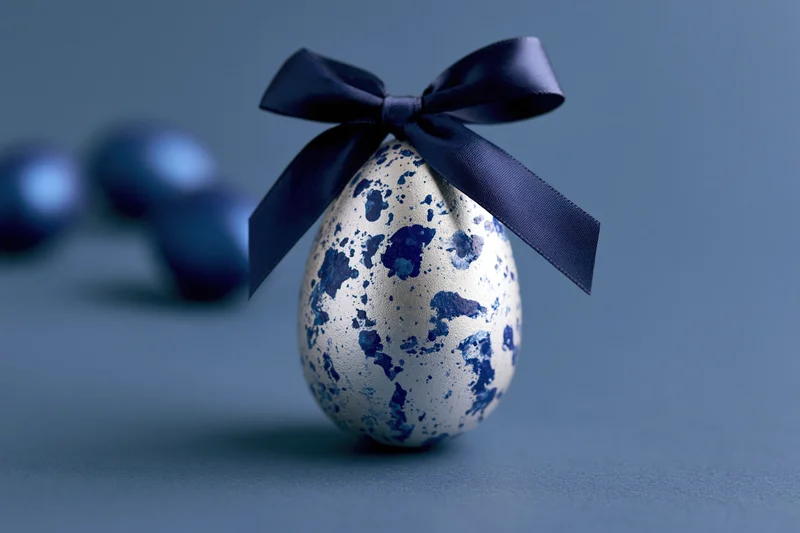 Silver easter egg with blue marbled paint splatters tied with a navy ribbon on a blue surface