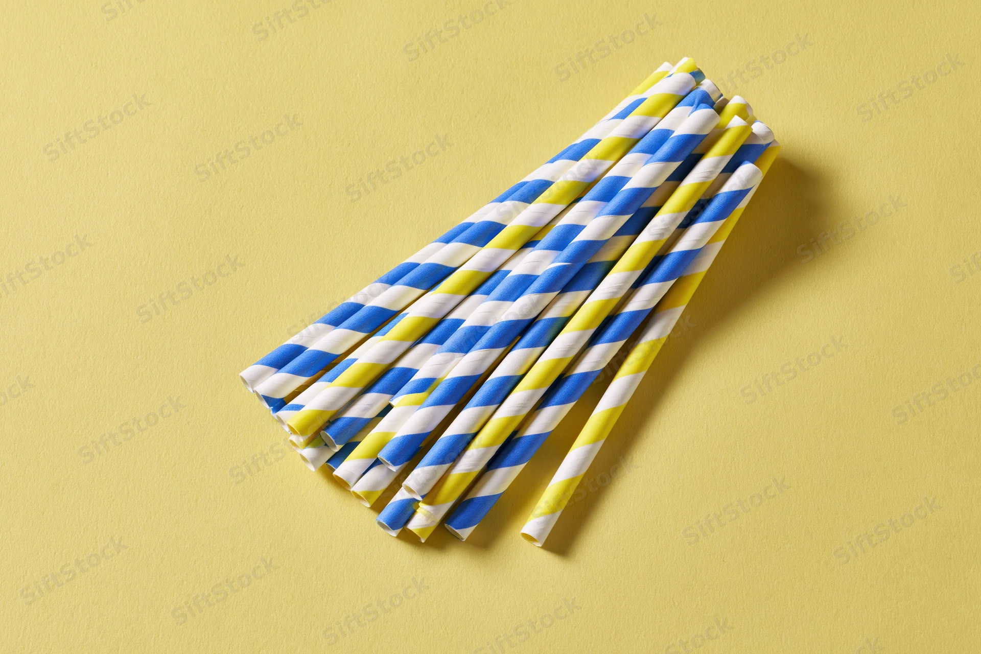 A bundle of blue, yellow and white striped paper drinking straws arranged diagonally on a yellow surface