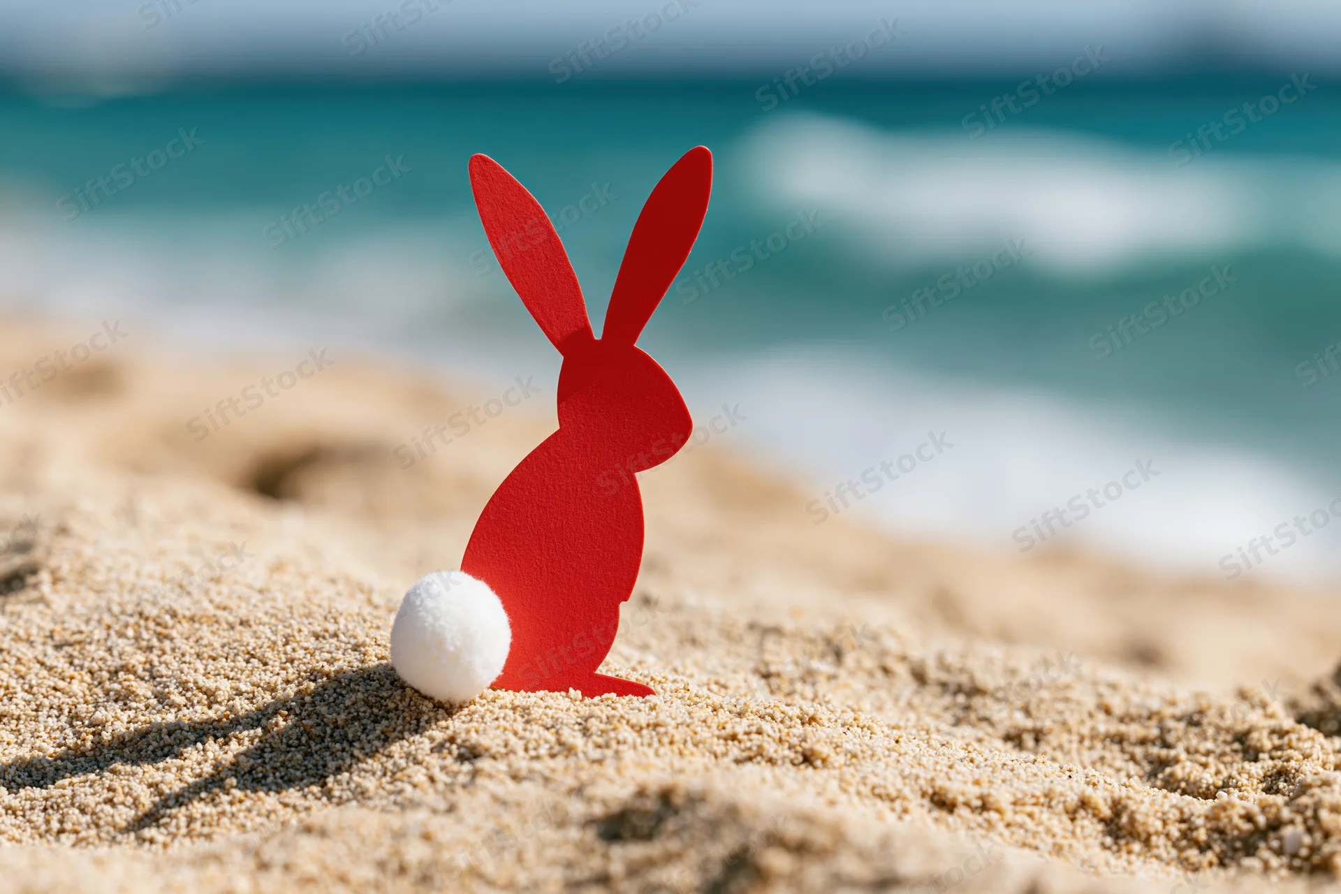 Red paper cutout easter bunny with white cotton tail standing on sandy beach near ocean waves