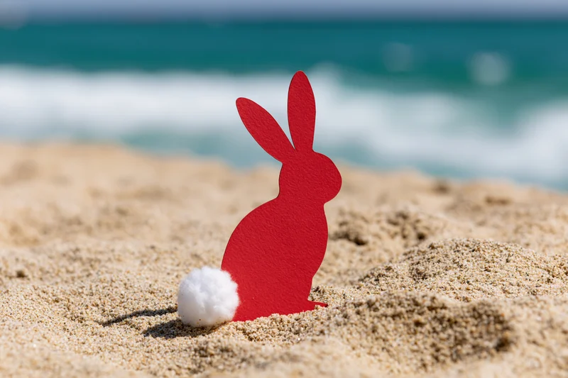 Red paper cutout easter bunny with white cotton tail placed on sandy beach near ocean waves