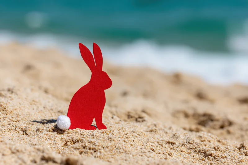 Red paper cutout easter bunny with white cotton tail sitting on sandy beach near ocean waves