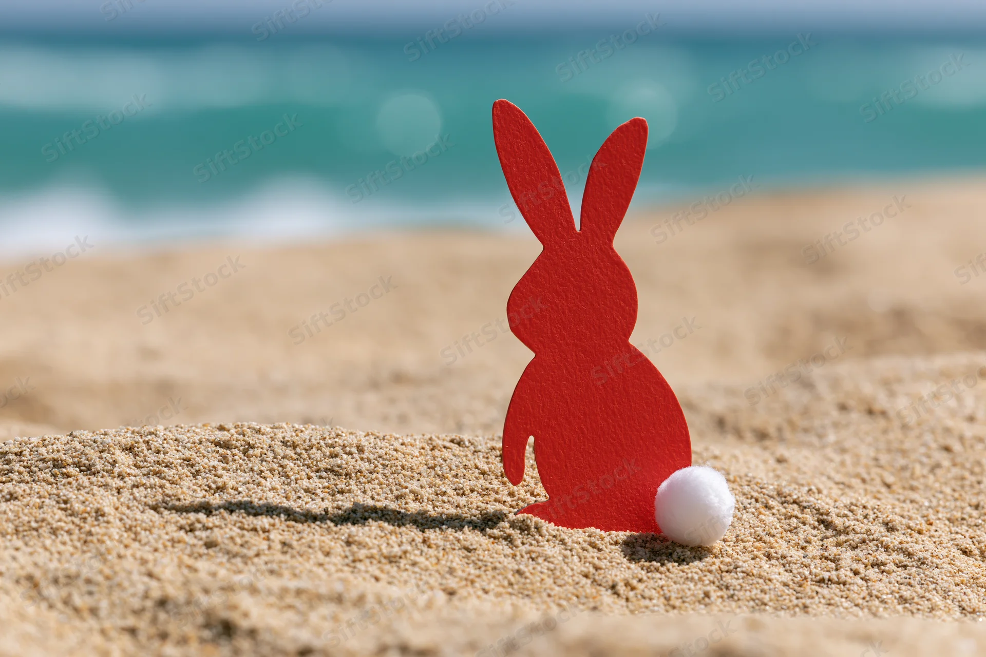 Red paper cutout easter bunny with white cotton tail standing on sandy beach near ocean waves