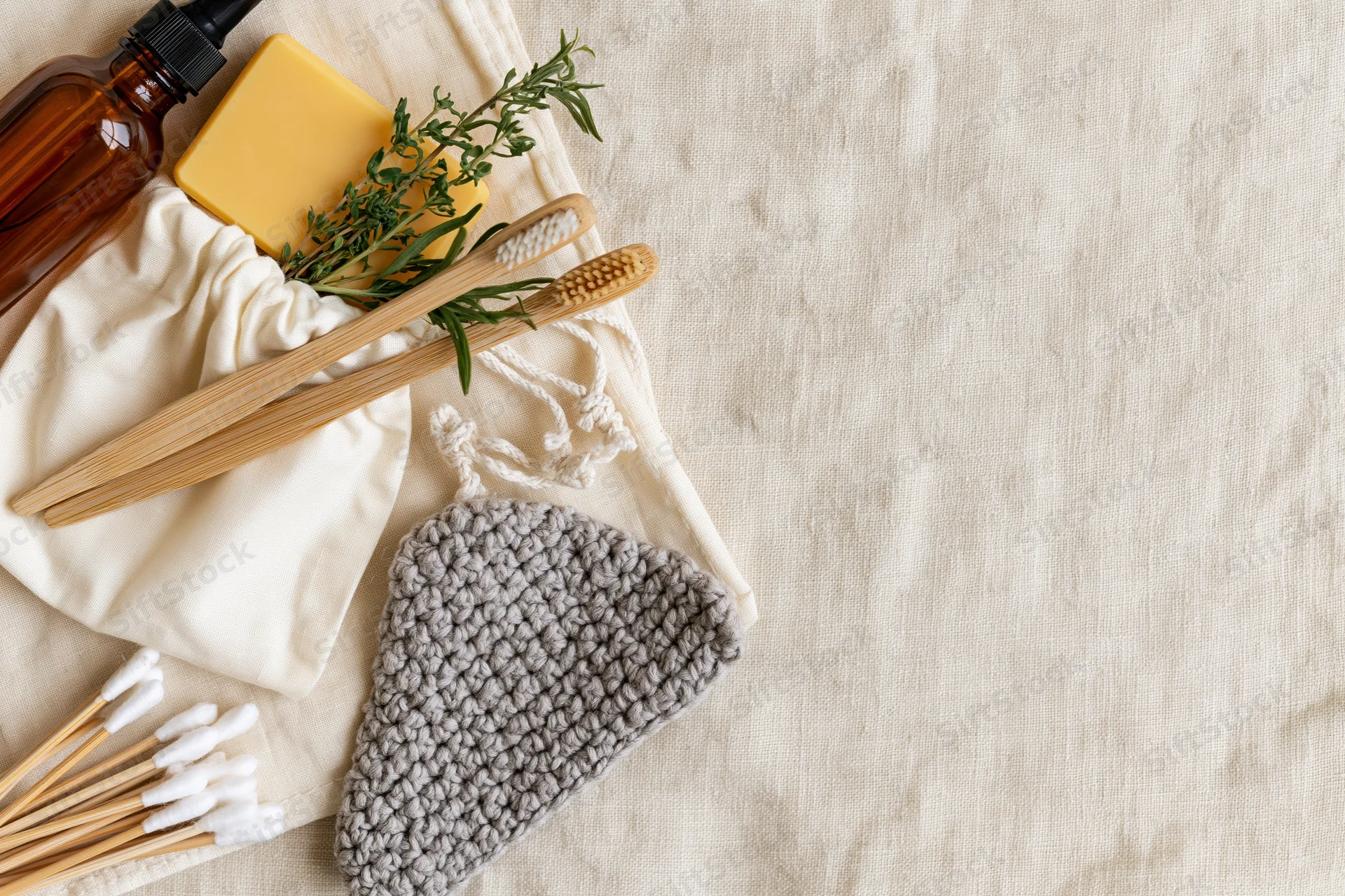 Bamboo toothbrushes and cotton buds spilling from a white drawstring pouch on a linen surface with natural skincare items and herbs