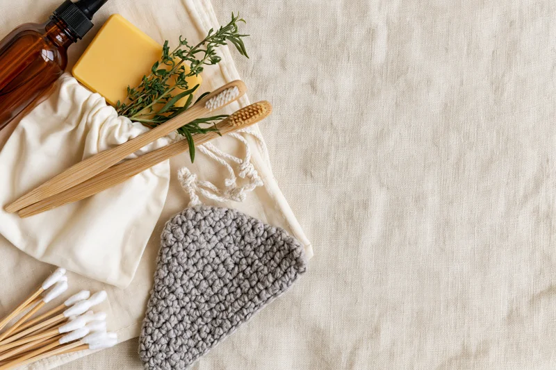 Bamboo toothbrushes and cotton buds spilling from a white drawstring pouch on a linen surface with natural skincare items and herbs