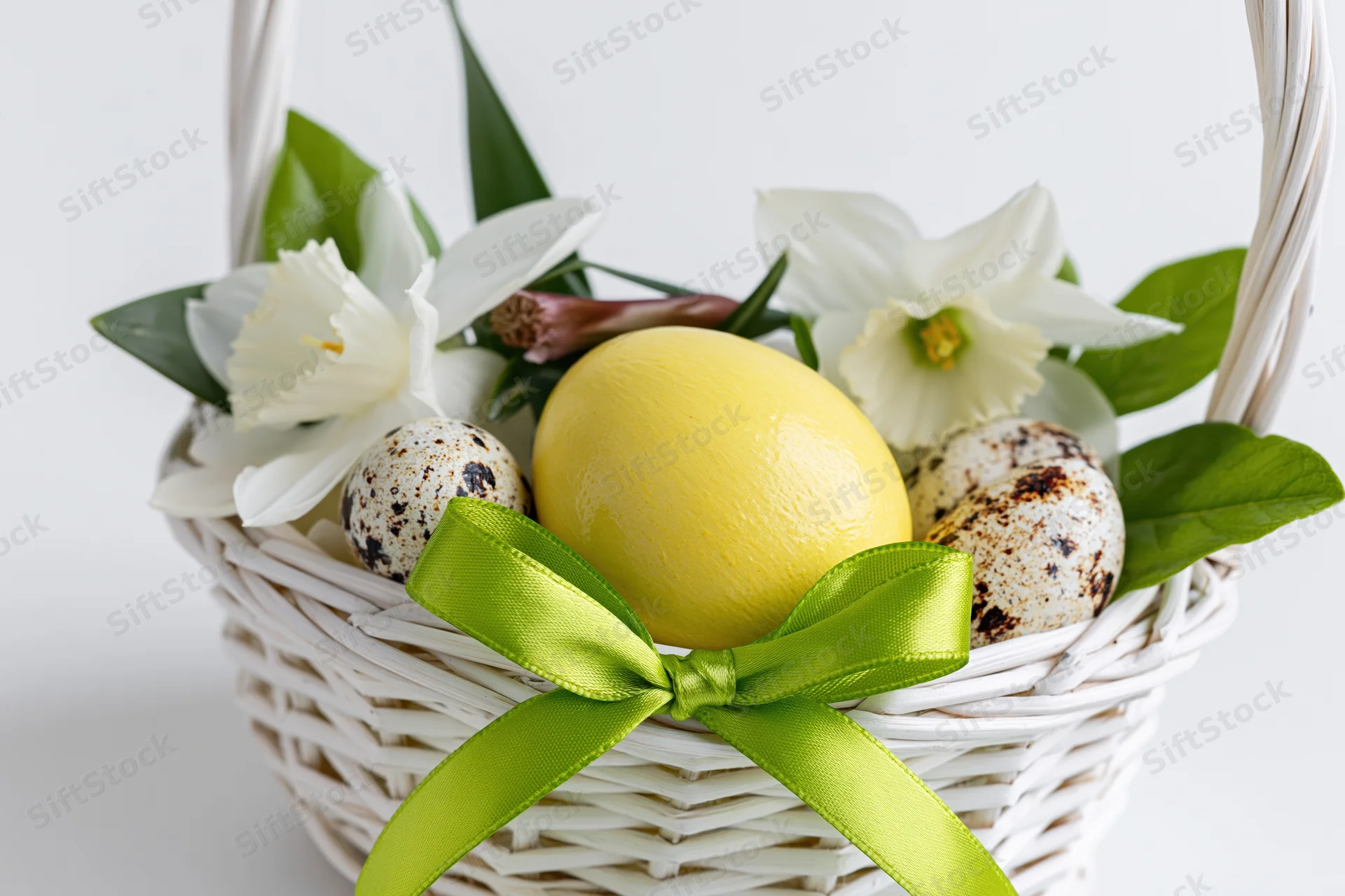 White wicker easter basket with yellow painted egg, speckled quail eggs, white daffodils and green ribbon bow arranged inside