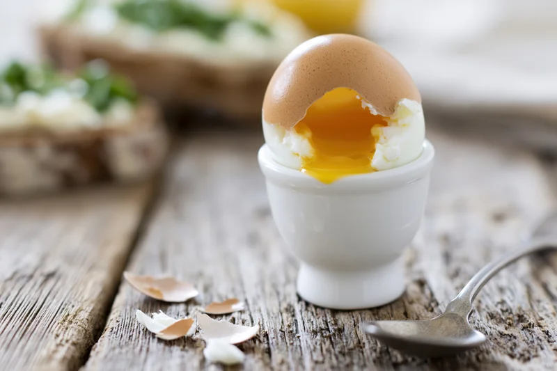 Soft-boiled egg cracked open in a white ceramic egg cup on a rustic wooden table with a spoon nearby