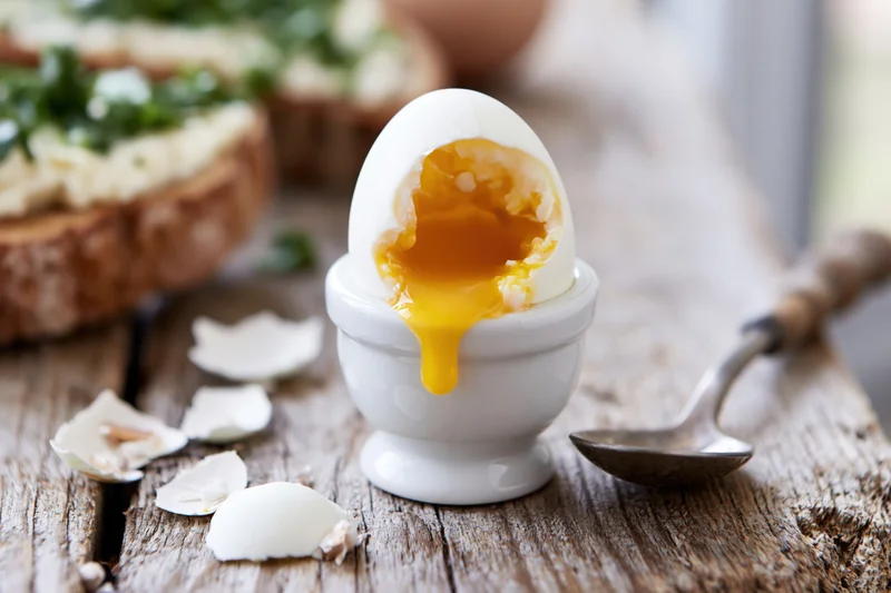 Soft-boiled egg cracked open in a white ceramic egg cup on a wooden table with toast and spoon nearby