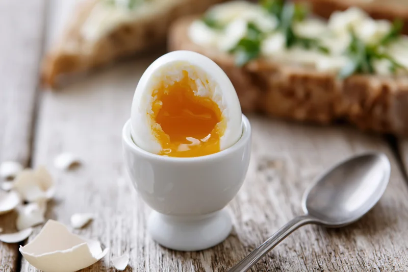 Soft-boiled egg cracked open in a white ceramic egg cup on a wooden table with toast and spoon nearby