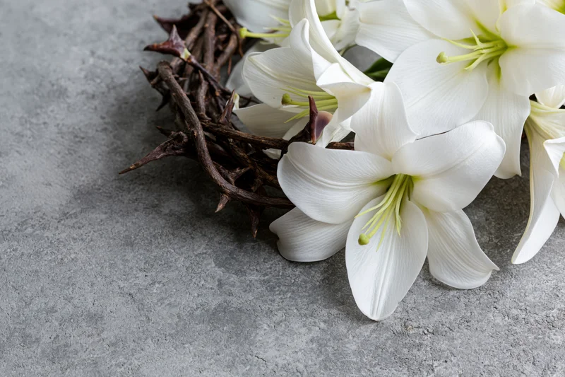 Woven crown of thorns resting beside white easter lilies on gray concrete surface