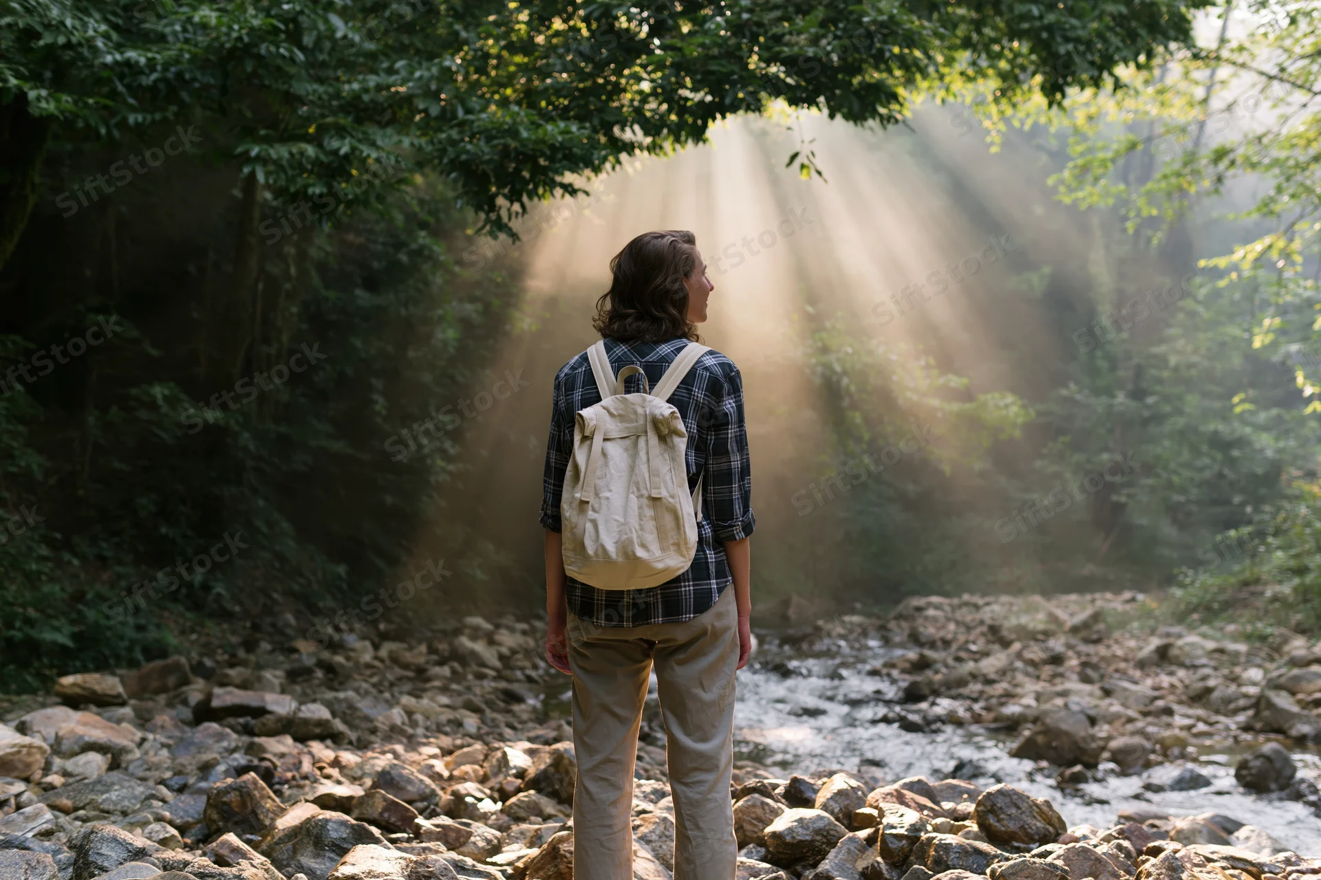 Woman with small backpack standing on rocky riverbed in forest with sunlight filtering through trees