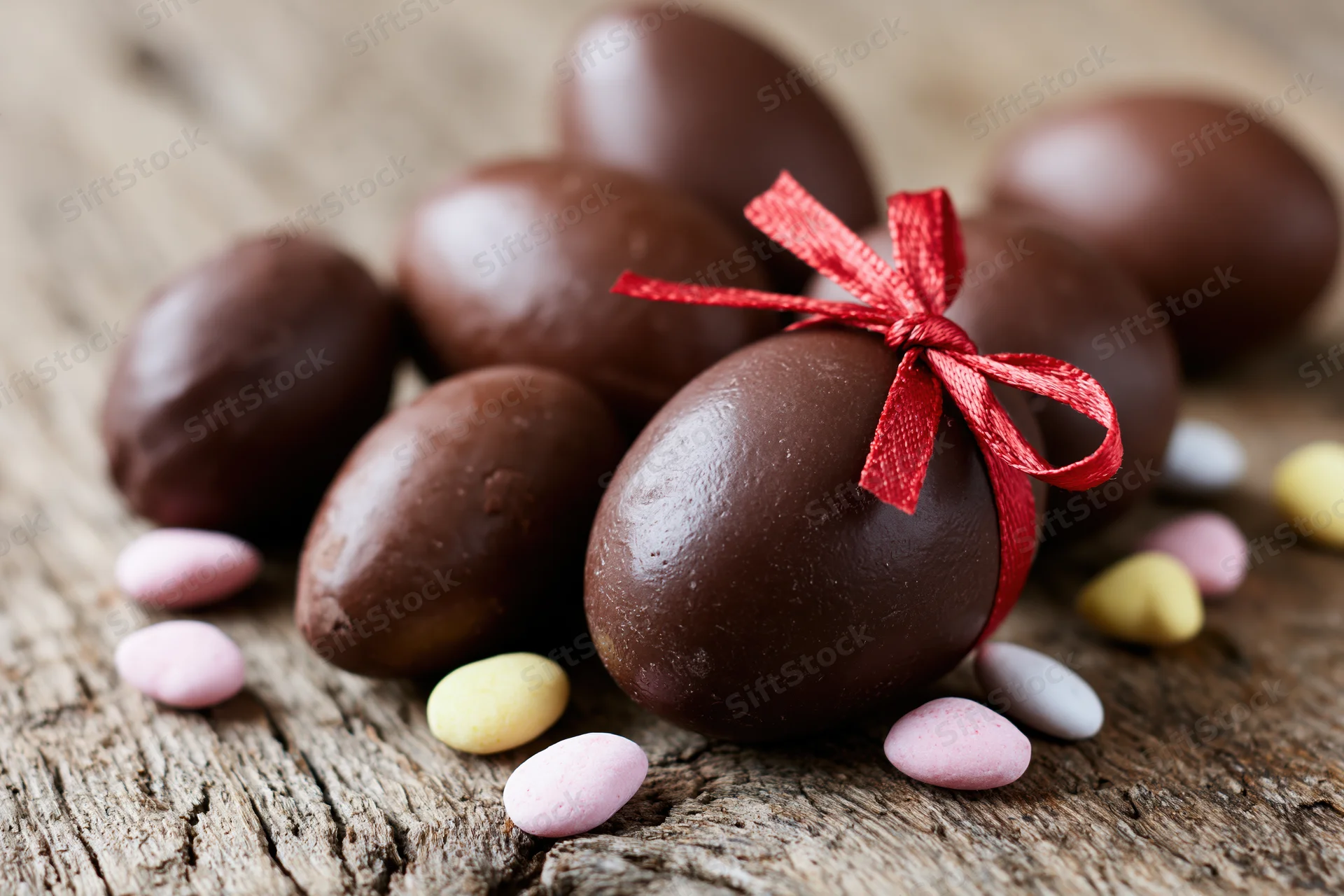 Chocolate easter eggs of varying sizes arranged on a rustic wooden surface with a red ribbon and scattered pastel candies