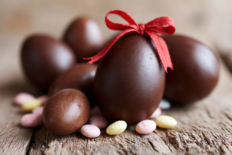 Assorted chocolate easter eggs of varying sizes arranged on a rustic wooden surface with a red ribbon and candy-coated chocolates scattered around