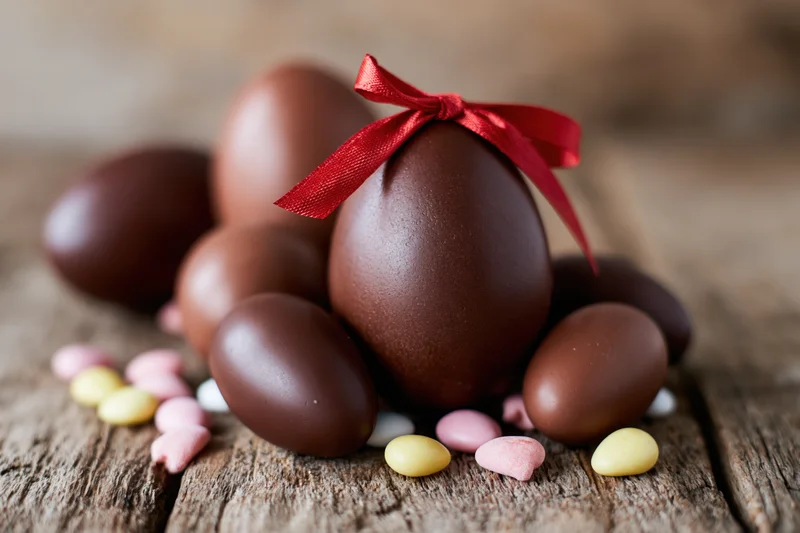 Assorted chocolate easter eggs of varying sizes arranged on a rustic wooden surface with a red ribbon and colorful candy hearts scattered around