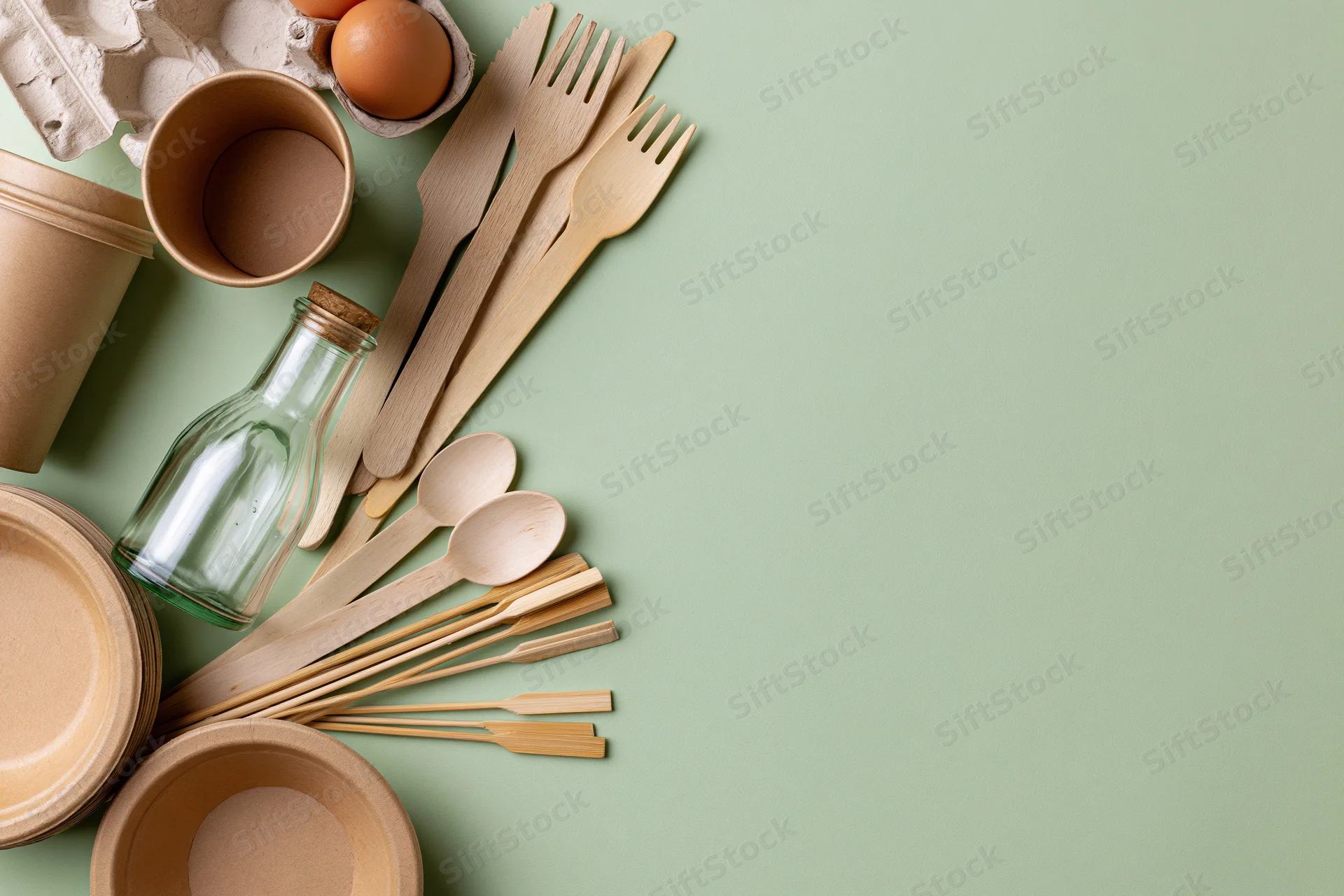 Eco-friendly disposable packaging arranged along the edge of a green surface with wooden cutlery, paper cups, glass bottle, and egg carton