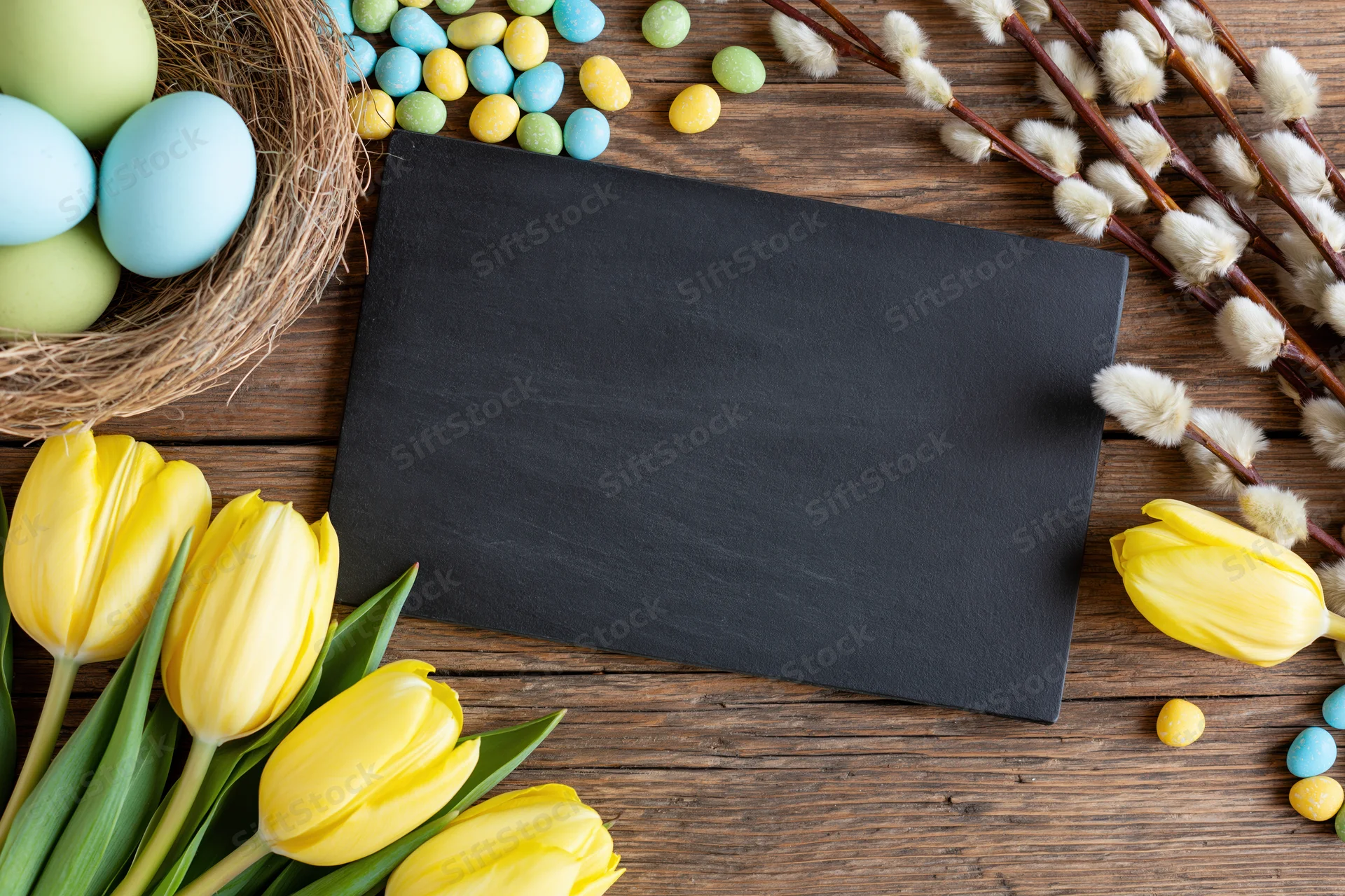 Blank black chalkboard card centered on rustic wooden table surrounded by easter decorations including yellow tulips, colorful eggs in a nest, and willow branches