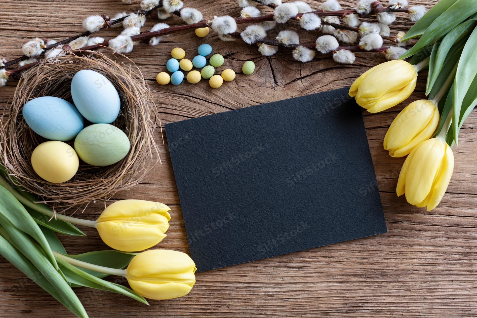 Blank black chalkboard card centered on rustic wooden table surrounded by easter eggs in a nest, yellow tulips, and willow branches
