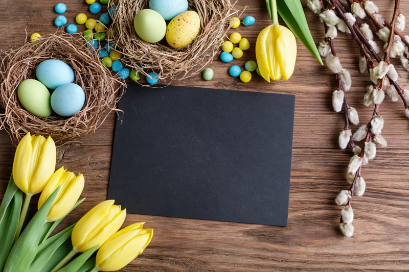 Blank black chalkboard card centered on rustic wooden table surrounded by easter decorations including nests with eggs, yellow tulips and willow branches