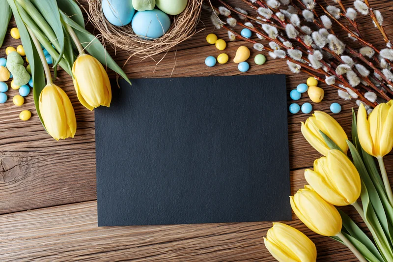 Blank black card centered on rustic wooden table surrounded by yellow tulips, easter eggs, and willow branches