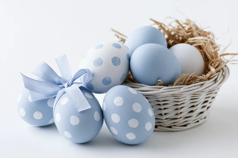 Blue and white painted easter eggs with polka dots arranged beside a wicker basket filled with straw on a plain white surface