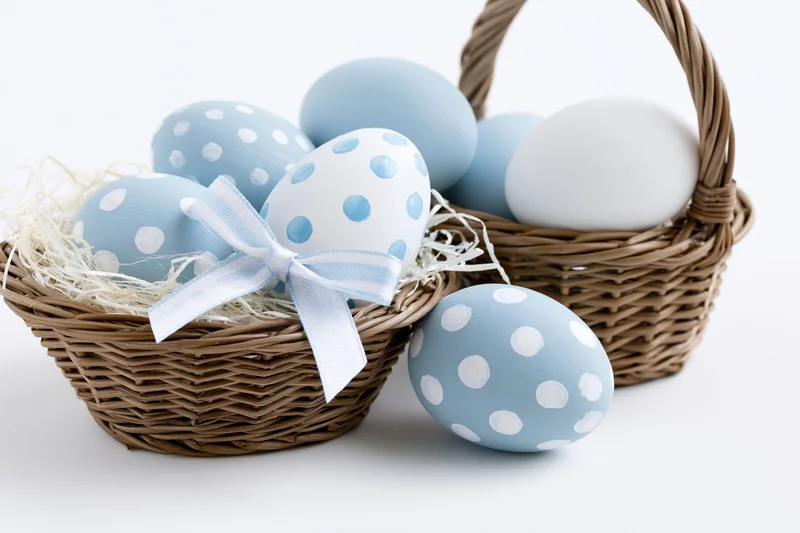 Blue and white polka-dotted painted easter eggs arranged in wicker baskets with straw and ribbon on white background