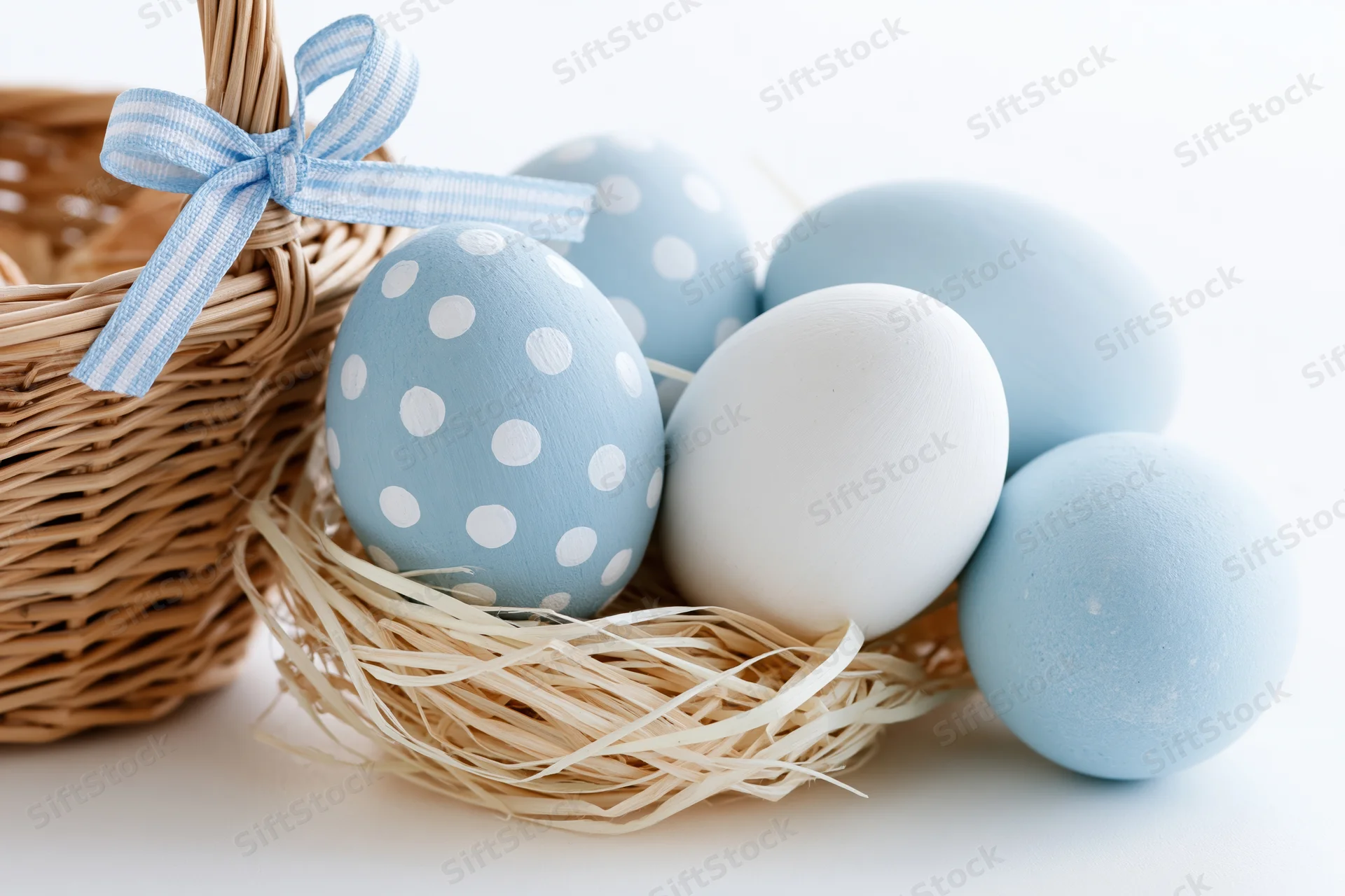 Painted blue and white easter eggs arranged in a straw nest beside a wicker basket with a checkered ribbon bow