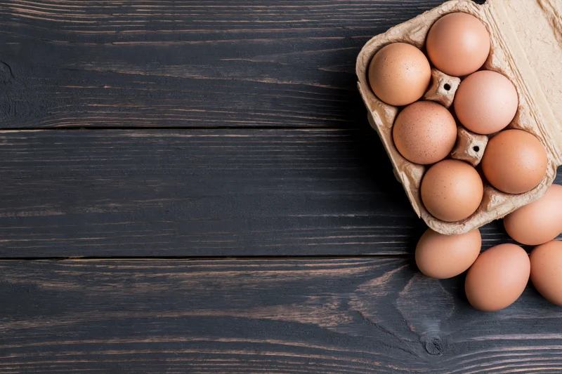 Brown chicken eggs in an open cardboard carton on a dark wooden table with some spilled out