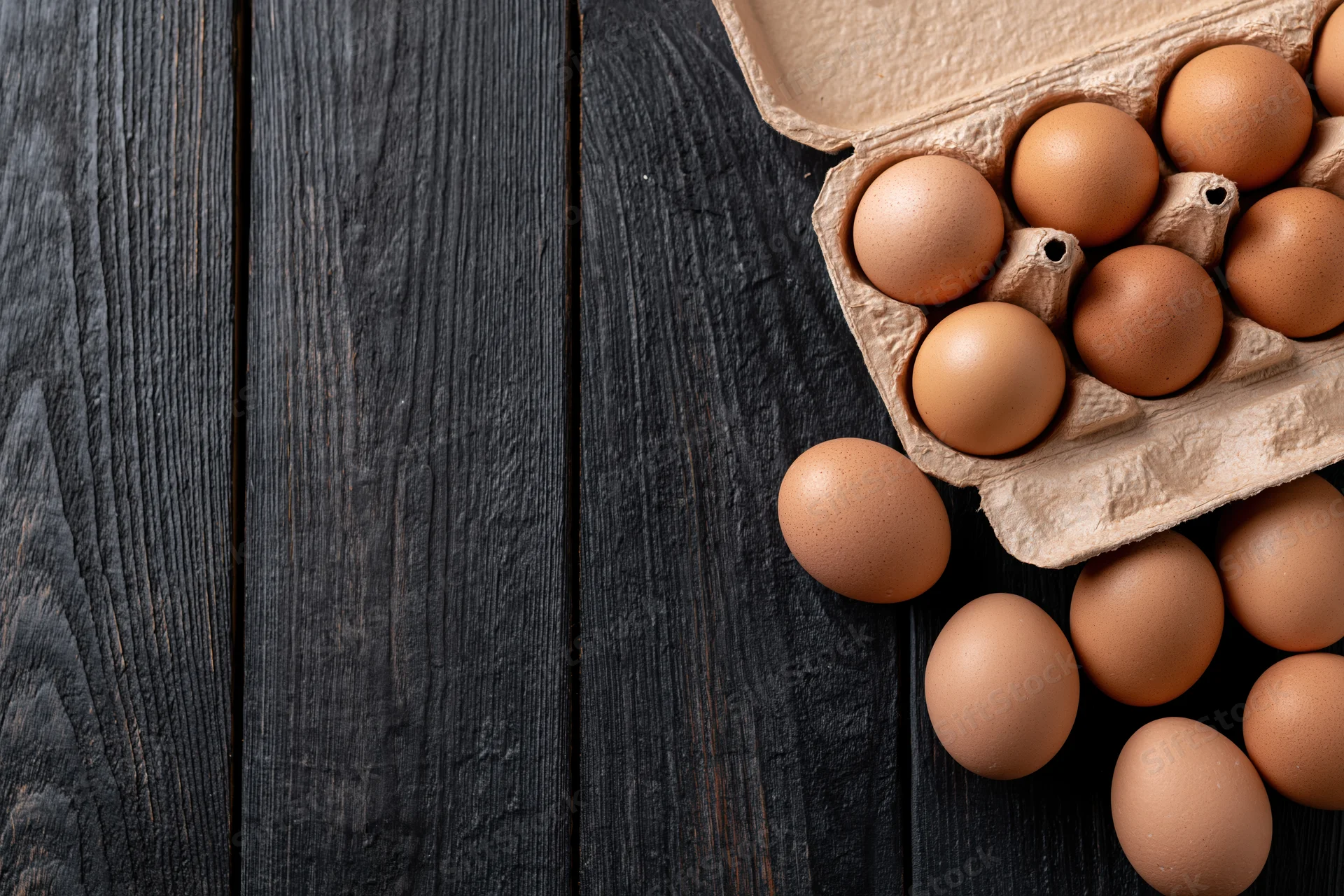 Brown chicken eggs in an open cardboard carton on a dark wooden surface with some eggs spilled beside it
