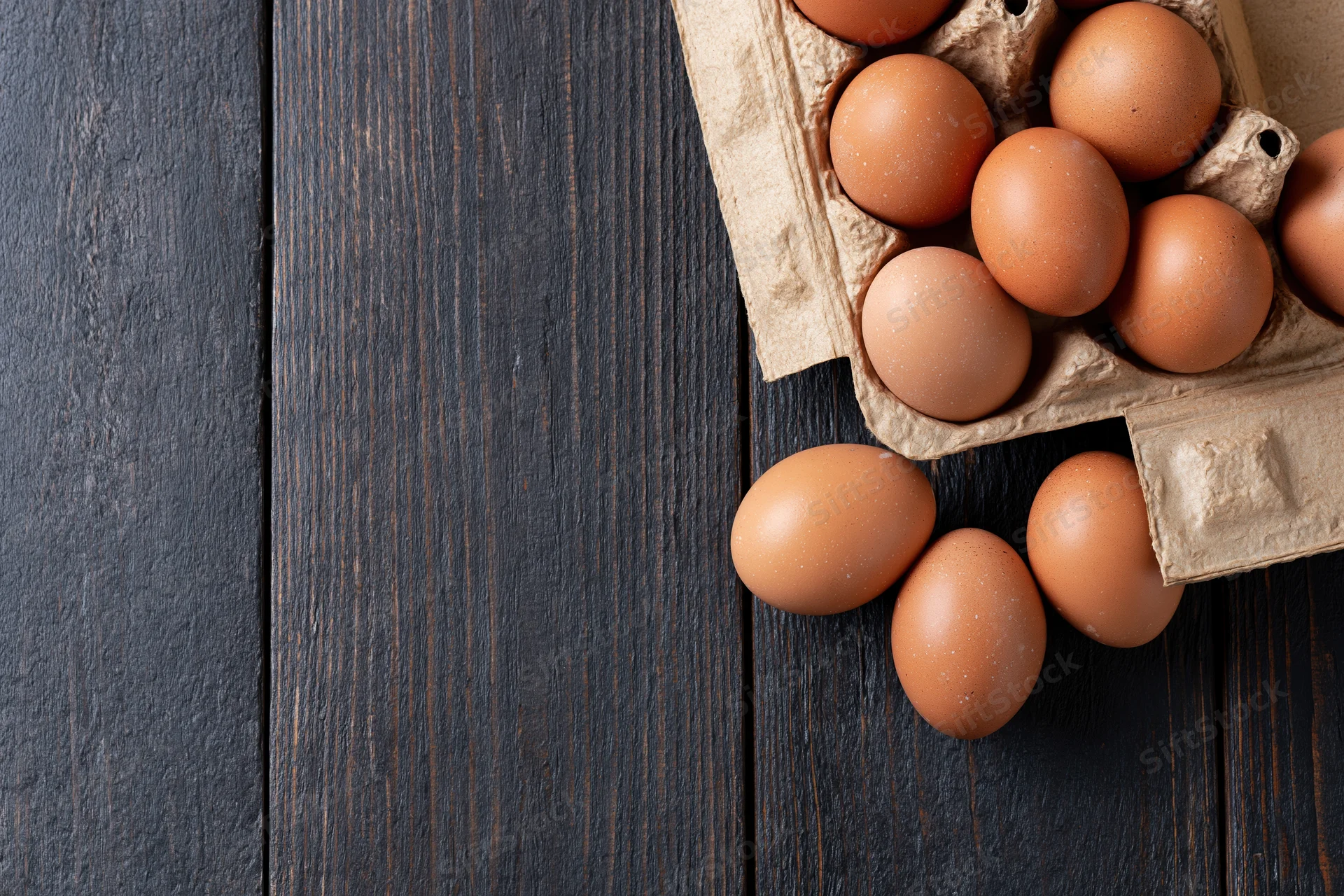 Brown chicken eggs in an open cardboard carton on dark wooden surface with some spilled out