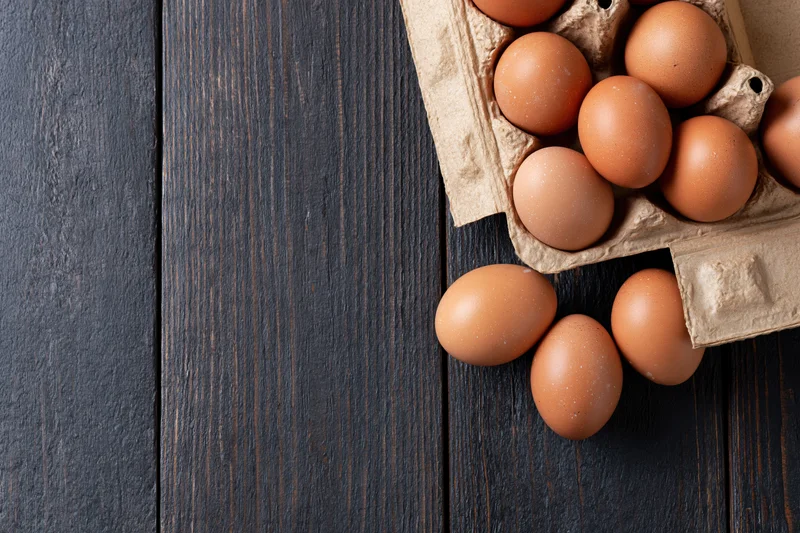 Brown chicken eggs in an open cardboard carton on dark wooden surface with some spilled out