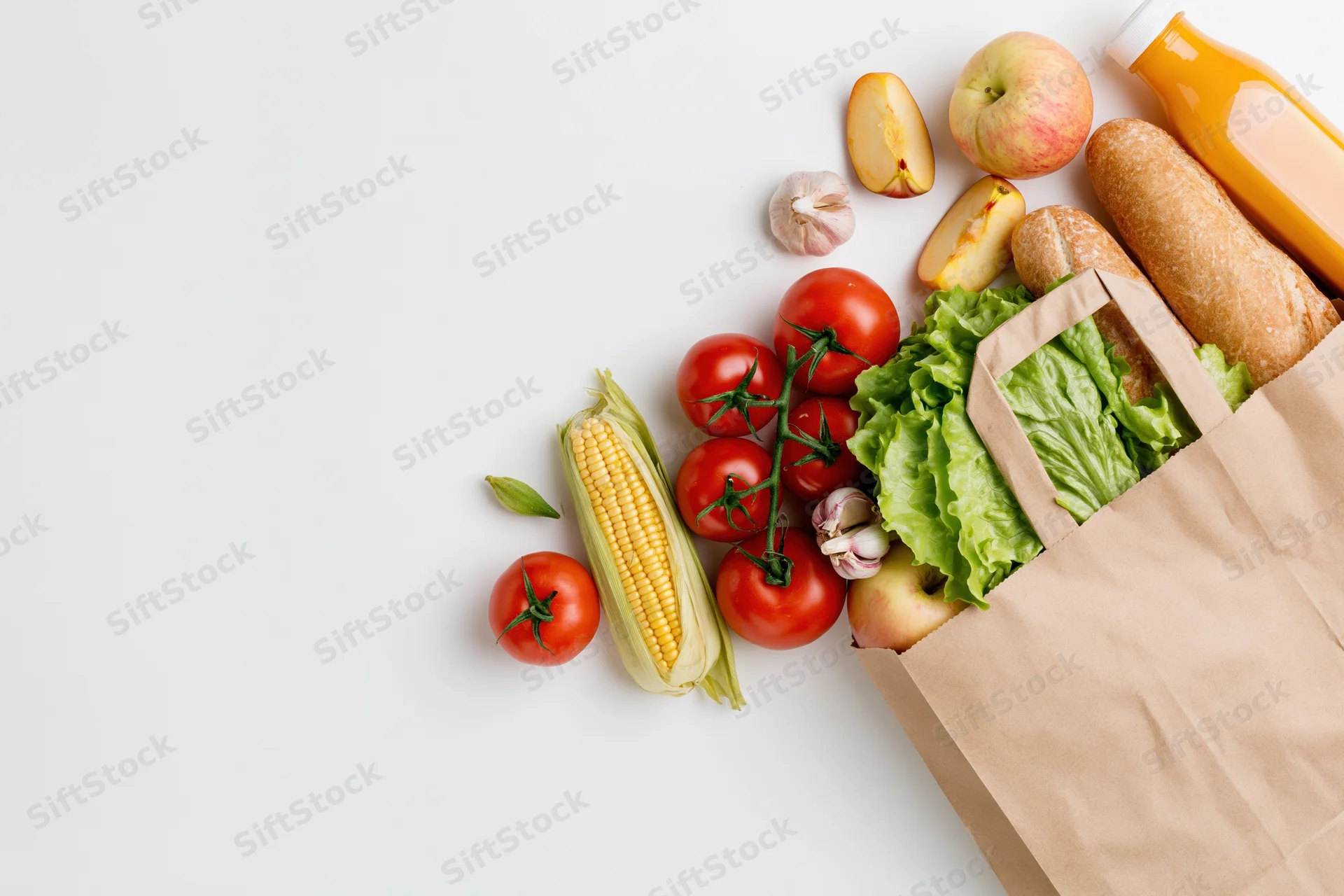 Brown paper grocery bag tipped on its side spilling fresh vegetables and fruits onto white surface