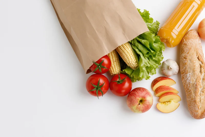 Brown paper grocery bag tipped over spilling fresh vegetables and fruits on white surface