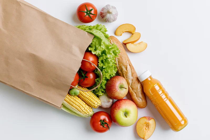 Brown paper grocery bag tipped over spilling fresh vegetables and fruits on white surface