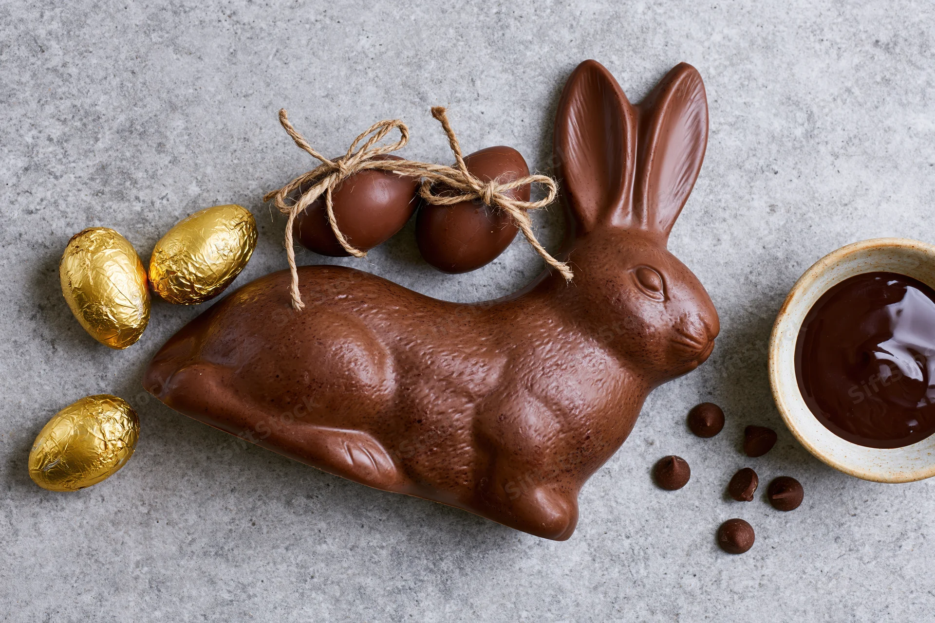 Chocolate easter bunny figurine lying on its side next to golden foil-wrapped eggs and a bowl of melted chocolate on a gray surface
