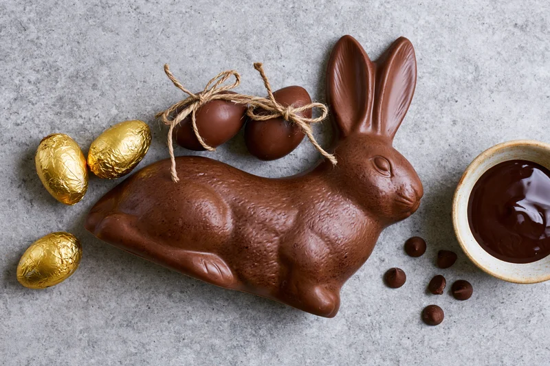 Chocolate easter bunny figurine lying on its side next to golden foil-wrapped eggs and a bowl of melted chocolate on a gray surface