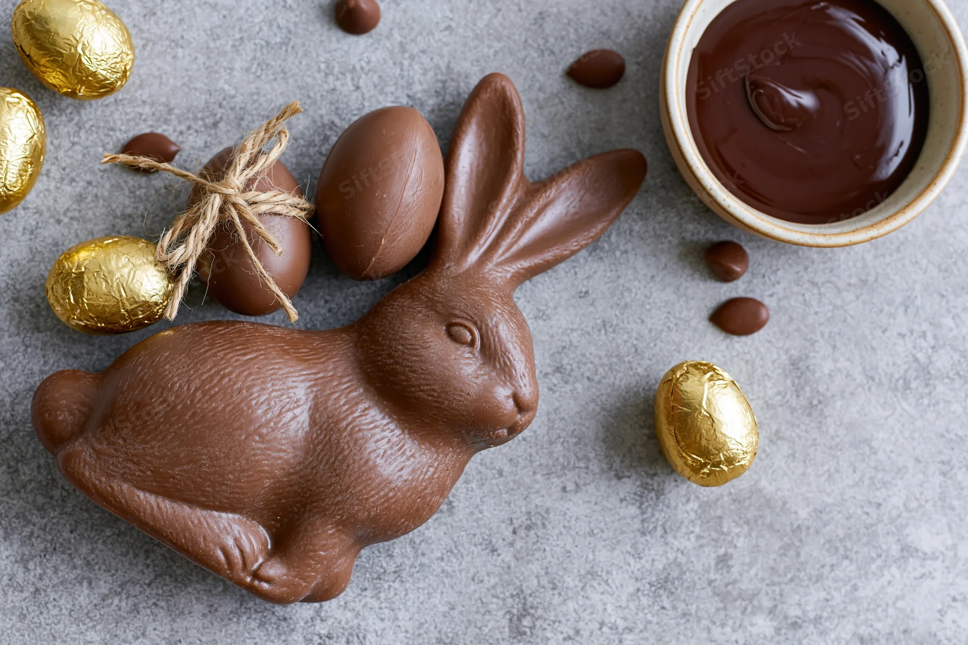 Chocolate easter bunny figurine lying on its side next to molten chocolate and golden foil-wrapped eggs on a gray stone surface