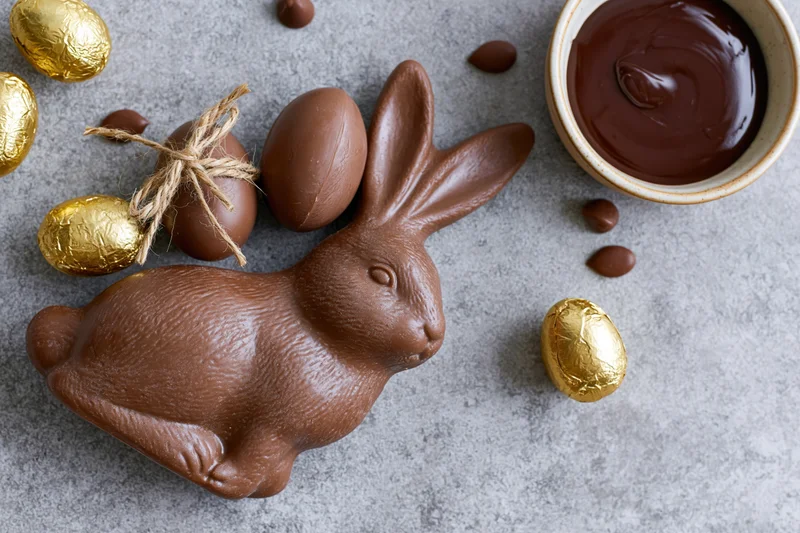 Chocolate easter bunny figurine lying on its side next to molten chocolate and golden foil-wrapped eggs on a gray stone surface