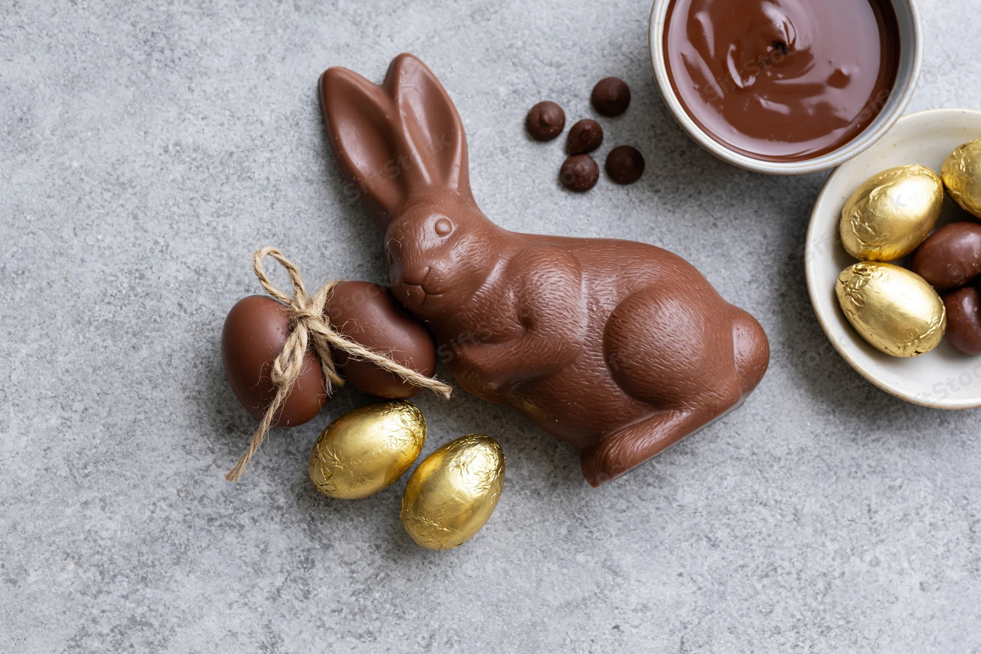 Chocolate easter bunny figurine lying on its side next to golden foil-wrapped eggs and a bowl of melted chocolate on a gray stone surface