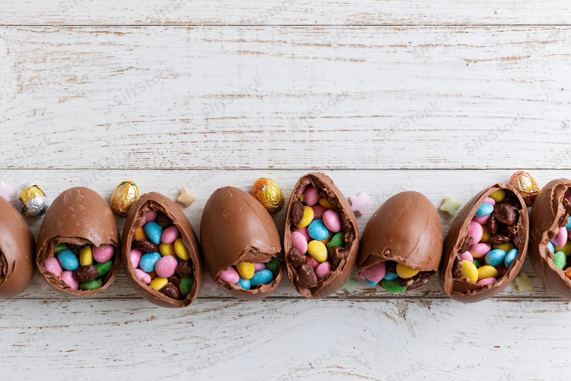 Cracked chocolate easter eggs filled with colorful candy arranged in a row on a white wooden surface