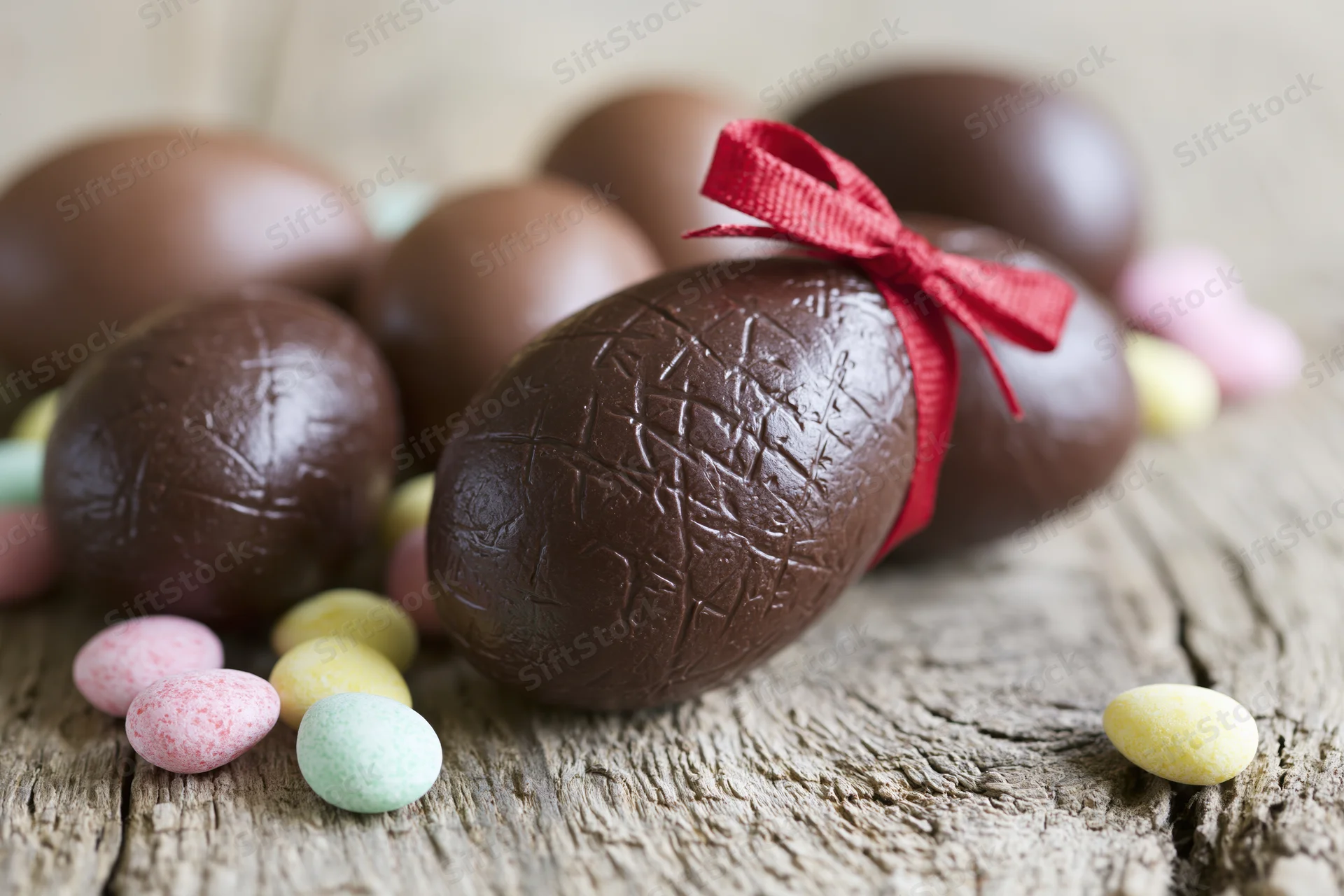Chocolate easter eggs of various sizes arranged on a weathered wooden surface with a red ribbon and small colorful candies scattered around
