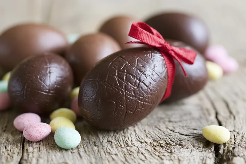 Chocolate easter eggs of various sizes arranged on a weathered wooden surface with a red ribbon and small colorful candies scattered around