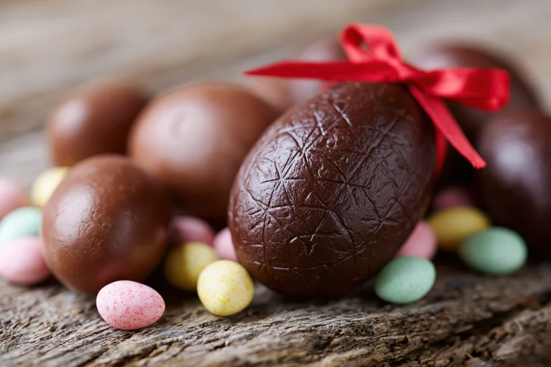 Chocolate easter eggs of various sizes arranged on a weathered wooden surface with a red ribbon and colorful candies scattered around