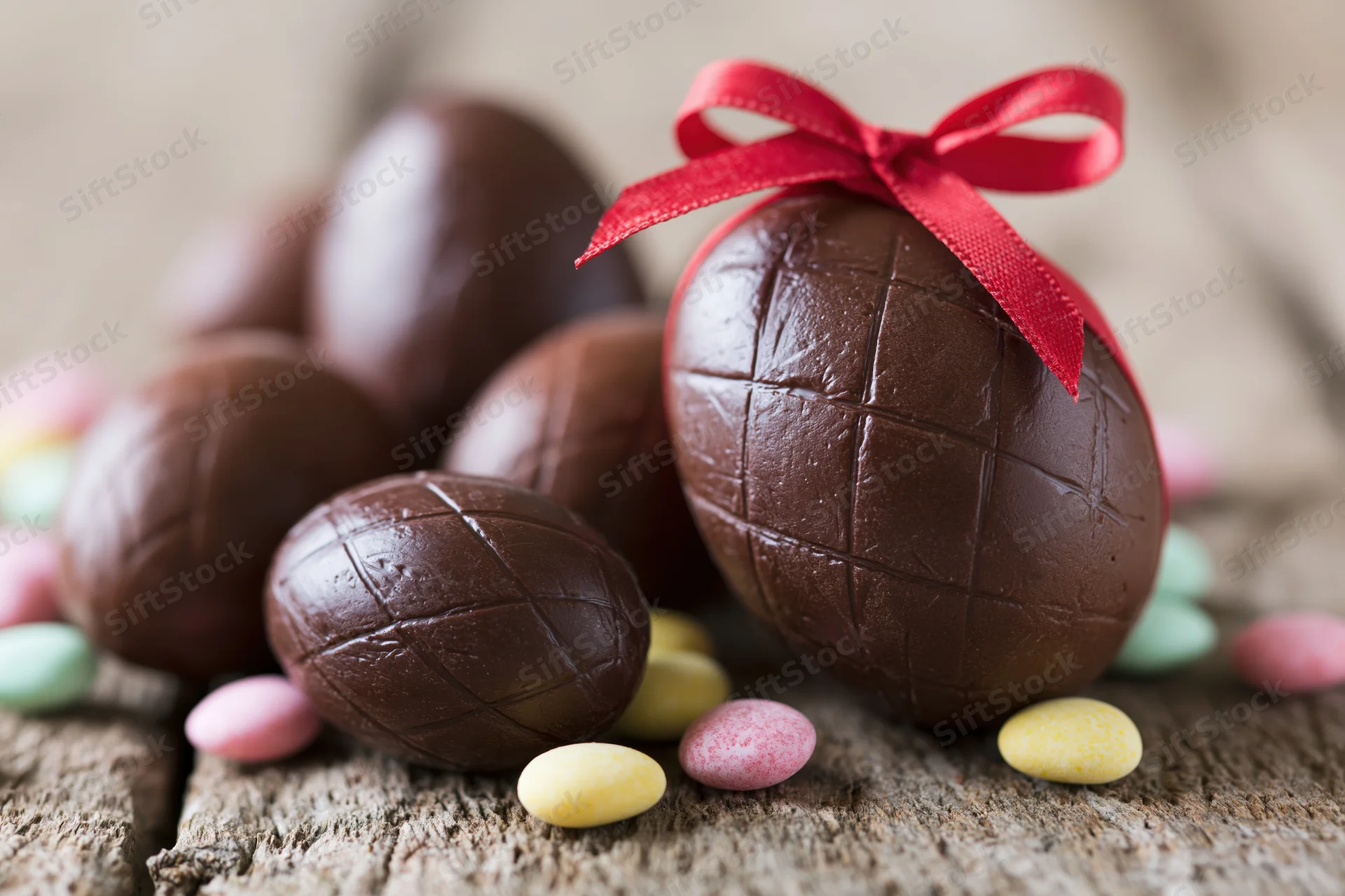 Chocolate easter eggs of various sizes arranged on a weathered wooden surface with a red ribbon and colorful candies scattered around