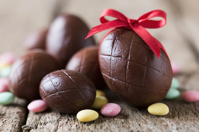 Chocolate easter eggs of various sizes arranged on a weathered wooden surface with a red ribbon and colorful candies scattered around