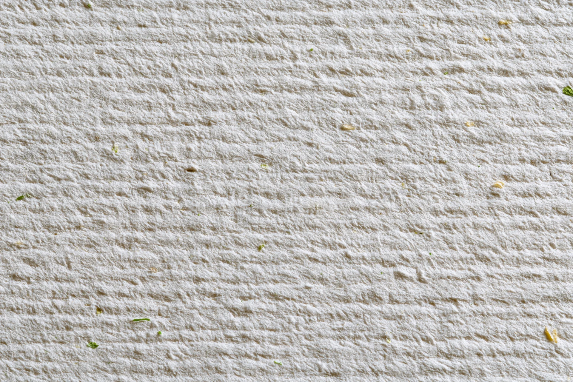 Close-up flat lay of off-white recycled paper with visible fibers and green plant fragments scattered across its textured surface