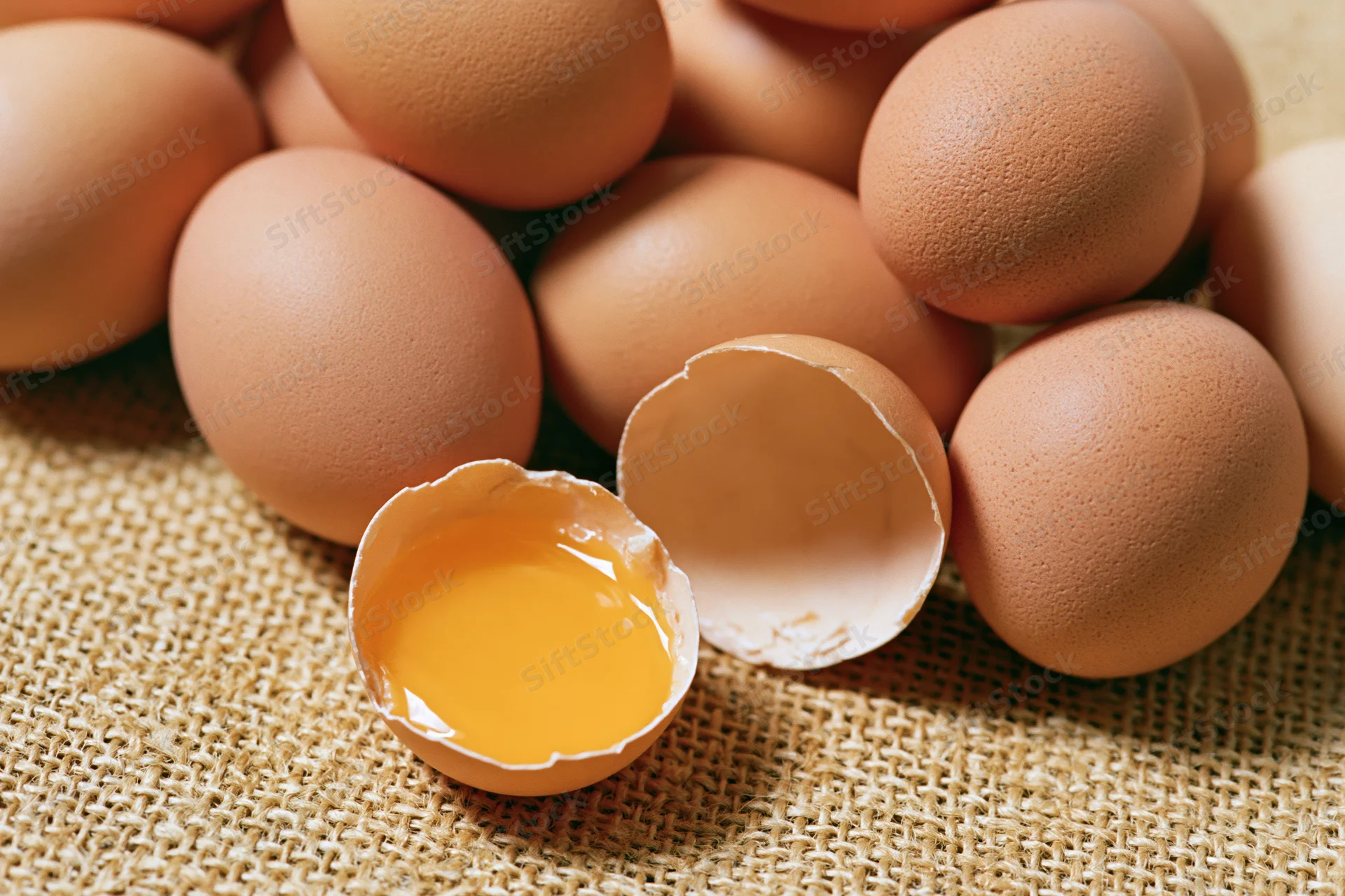 Cracked brown chicken egg split in two halves with yolk visible on burlap surface surrounded by whole eggs