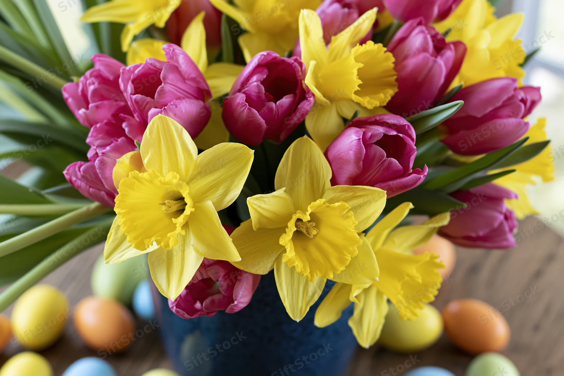 Spring flower bouquet with yellow daffodils and pink tulips in blue vase on wooden table surrounded by easter eggs