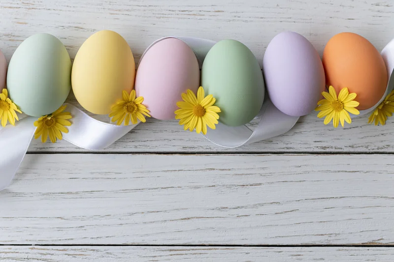 Pastel easter eggs arranged in a row on a white wooden surface with yellow daisies and a white ribbon
