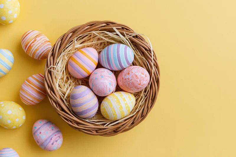 Colorful hand-painted easter eggs arranged in a wicker basket with straw on a yellow background
