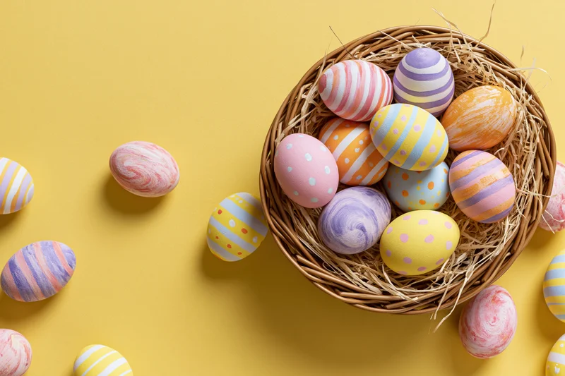 Colorful hand-painted easter eggs arranged in a wicker basket with straw on yellow background