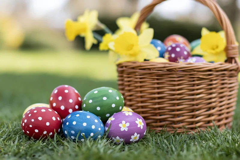 Colorful hand-painted easter eggs arranged on green grass beside a wicker basket with daffodils